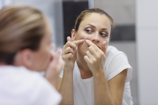 Jeune femme à la maison se regarde dans le miroir et fait sortir un bouton. Soins des peaux à problèmes, nettoyage des mains