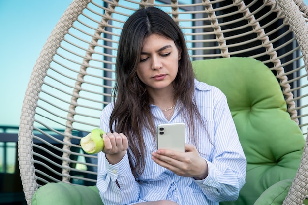 Une jeune femme mange une pomme avec un smartphone dans les mains