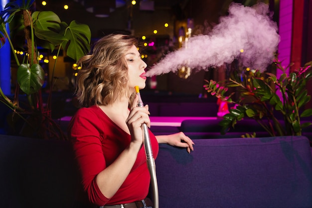 Jeune femme à la robe rouge fume un hookan. La discothèque ou le bar fument la chicha.