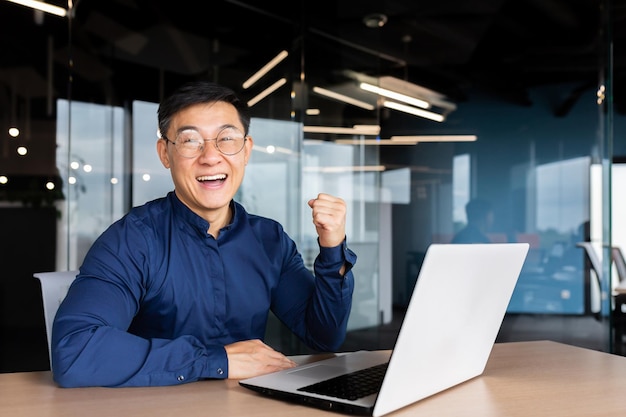 Photo jeune homme d'affaires asiatique prospère et heureux, il est assis au bureau sur l'ordinateur portable et se réjouit d'une victoire