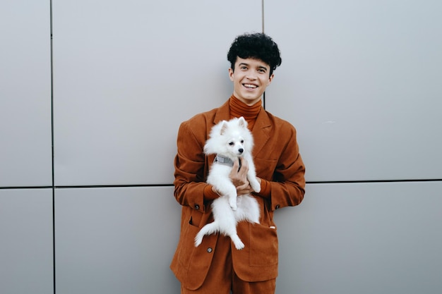 Photo un jeune homme en costume brun tenant un petit chien blanc contre un mur gris il a une expression joyeuse