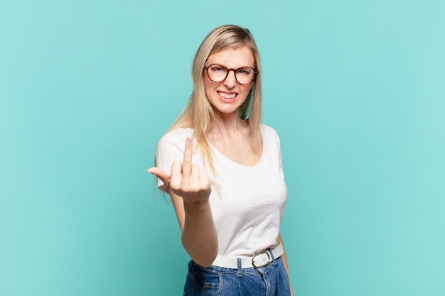 Jeune jolie femme blonde se sentant en colère, agacée, rebelle et agressive, renversant le majeur, ripostant