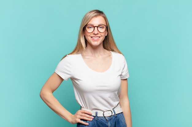 Jeune jolie femme blonde souriante joyeusement avec une main sur la hanche et une attitude confiante, positive, fière et amicale