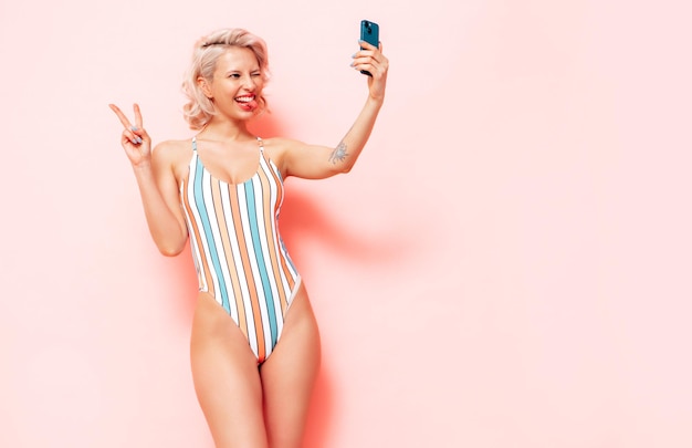 Jeune mannequin blonde souriante en maillot de bain d'été maillot de bain coloré Sexy femme insouciante s'amuser et devenir folle Femme posant près du mur rose en studio Enthousiaste et heureux Prendre selfie