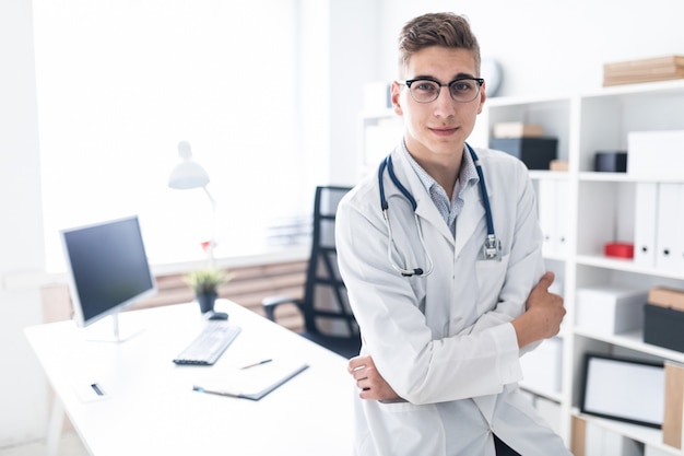 Un jeune médecin en robe blanche est appuyé sur la table du bureau.