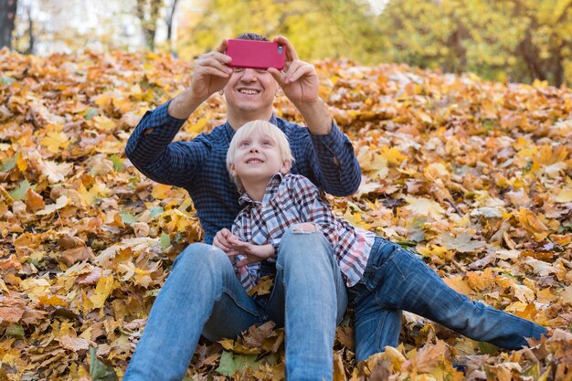 Jeune père et enfant mignon assis dans le parc d'automne sur les feuilles tombées. Papa et garçon font des photos pour téléphone.