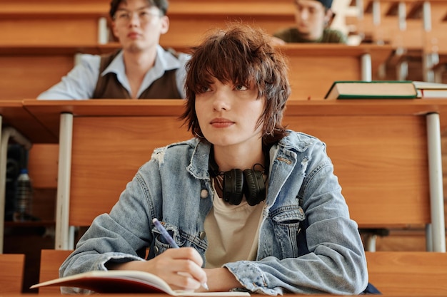 Photo jolie adolescente en veste en jean prenant des notes tout en écoutant le professeur