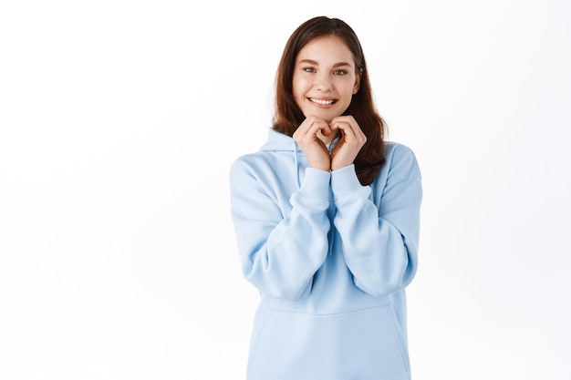 Jolie jeune femme en sweat à capuche, montrant le geste du cœur de la main et vous souriant, amour et signe comme, debout contre le mur blanc