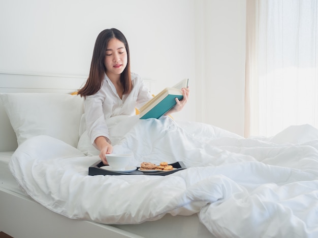 Livre de lecture de femme asiatique et de boire du café sur le lit le matin