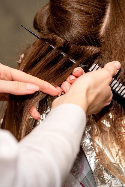 Les mains du coiffeur préparent les cheveux bruns pour la teinture avec un peigne et une feuille dans un salon de beauté en gros plan