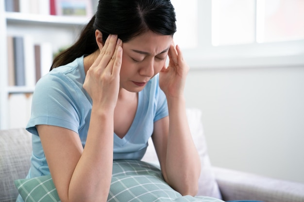 Photo maman japonaise femme souffrant de maux de tête assis sur un canapé tenant un oreiller avec un visage froncé. une femme asiatique malade à la maison qui se repose ne va pas travailler avec le stress de la tête qui fait mal. maladie femme à l'intérieur.