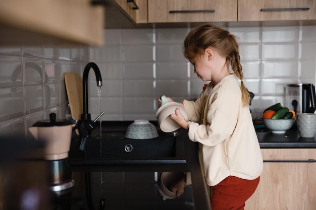 Un petit enfant avec un chiffon dans les mains se tient sur une chaise près de l'évier et lave la vaisselle Une petite fille aide sa mère à cuisiner et à nettoyer dans la cuisine