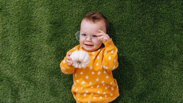 Un petit enfant heureux se trouve sur l'herbe verte dans un pull jaune et des lunettes tient une citrouille dans ses mains