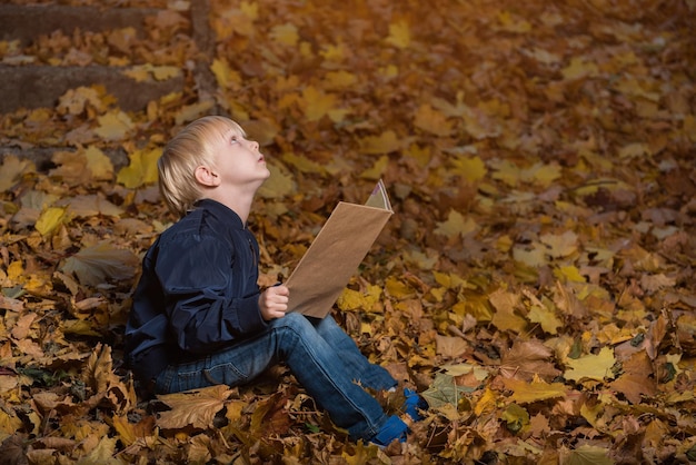 Petit garçon est assis dans la forêt sauvage dans les feuilles d'automne avec un livre dans les mains Livres pour enfants sur les miracles et la magie