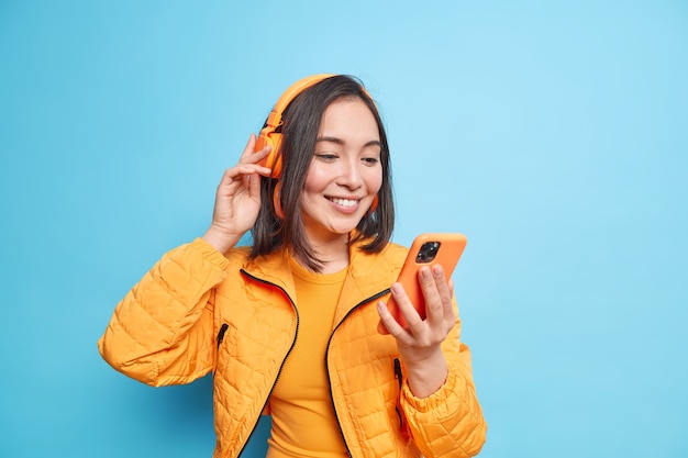 Une photo horizontale d'une jolie femme asiatique reçoit un message sur les réseaux sociaux concentré sur un smartphone écoute de la musique via des écouteurs sans fil tout en marchant porte une veste orange isolée sur un mur bleu