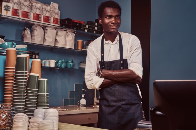 Photo portrait d'un beau barista africain debout avec les bras croisés au comptoir d'un café branché.