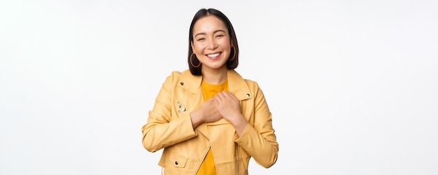 Portrait d'une belle fille asiatique souriante regardant avec appréciation merci geste tenant la main sur le cœur flatté debout sur fond blanc