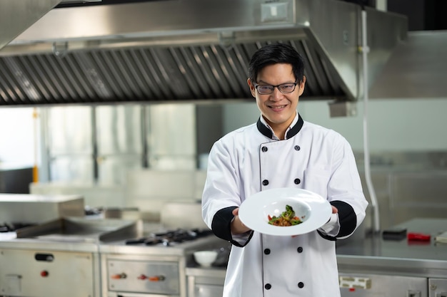 Photo portrait de chef asiatique thaïlandais regardant la caméra avec un coup de poing et montrant de la nourriture dans une assiette blanche cours de cuisine salle de classe de cuisine école de cuisine leçons de cuisine dans une école de cuisson