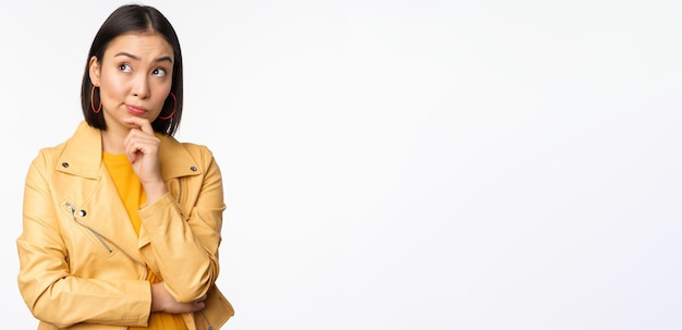 Portrait d'une femme coréenne élégante en veste jaune souriante réfléchie et regardant le logo ou la publicité debout sur fond blanc