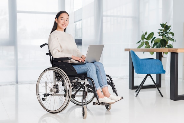 Portrait d&#39;une jeune femme souriante assise sur un fauteuil roulant, regardant la caméra avec un ordinateur portable sur ses genoux