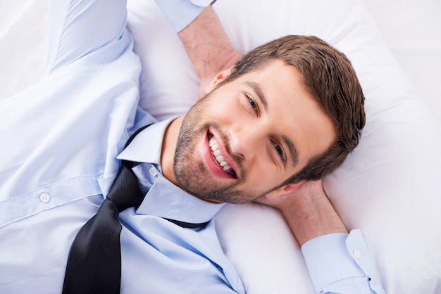 Rêveur heureux. Vue de dessus du beau jeune homme en chemise et cravate tenant les mains derrière la tête et souriant en position couchée dans son lit