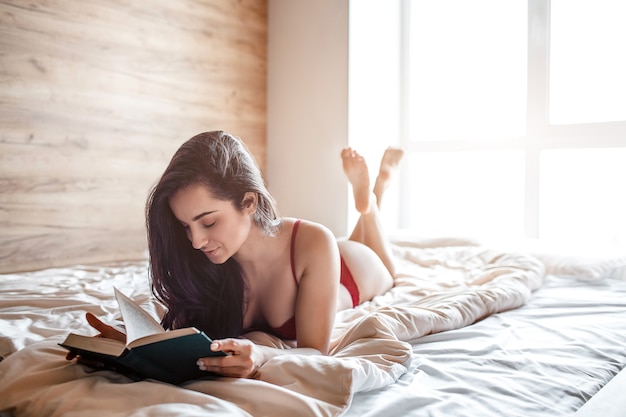 Séduisante jeune femme brune nue sur le lit le matin. Modèle sexy chaud couché et livre de lecture. Calme et paisible. Joyeux modèle bien construit. Personne attirante.