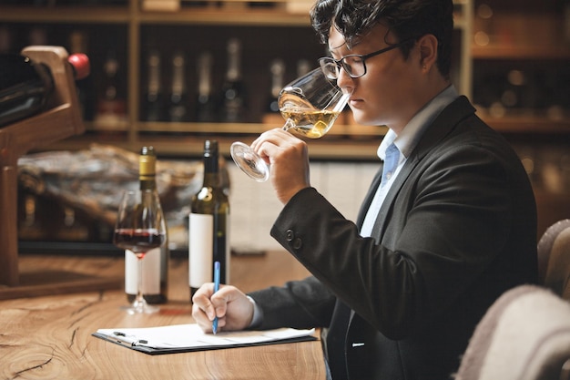 Photo sommelier tenant un verre de vin par l'odeur et écrivant des notes assis dans la cave