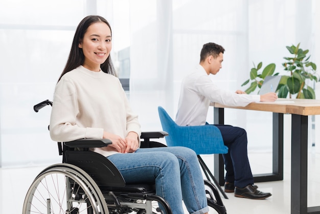 Souriant handicapée jeune femme assise sur un fauteuil roulant, regardant la caméra devant l&#39;homme travaillant sur un ordinateur portable