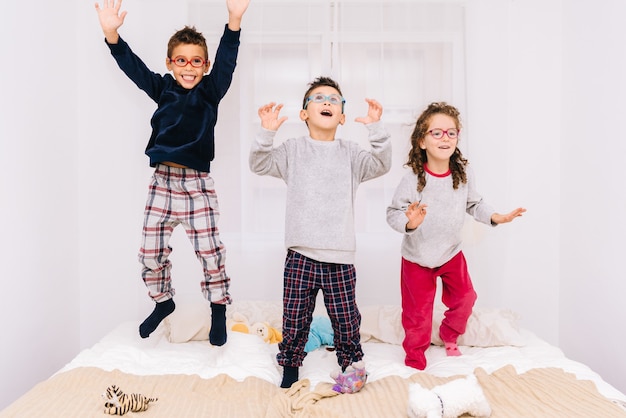 Trois enfants joyeux avec des lunettes sautant et jouant sur le lit