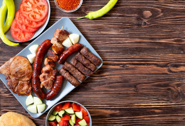 Viande grillée traditionnelle serbe et balkanique appelée mesano meso. Barbecue balkanique (rostilj) servi avec salade serbe, piments forts, pain, tomate, oignons et paprika en poudre. Fond en bois. Vue de dessus