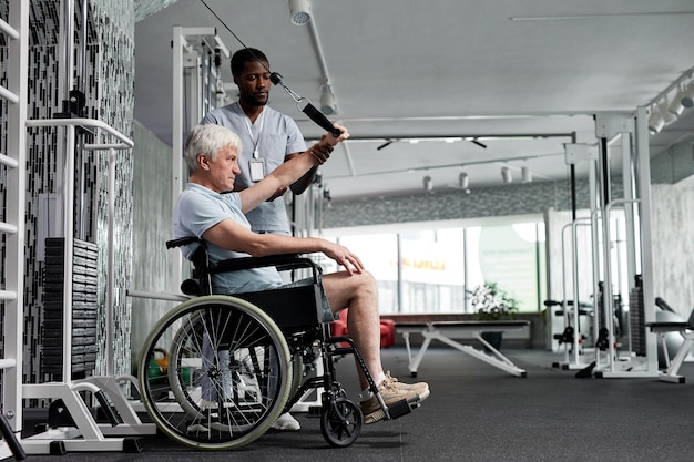 Vue latérale portrait d'un homme âgé utilisant un fauteuil roulant dans une salle de sport et faisant des exercices de rééducation avec le cul