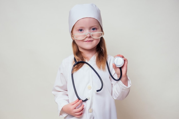 Adorable child girl uniformed as doctor wearing doctor's cap and glasses with stethoscope on white background.Happy kid little female doctor career guidance dreaming about future profession