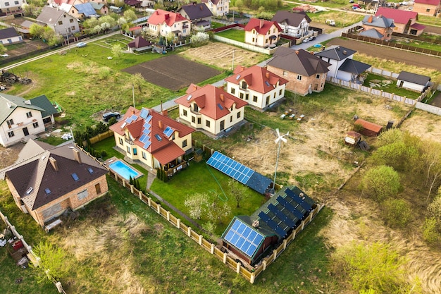 Aerial view of a private house with green grass covered yard, solar panels on roof, swimming pool with blue water and wind turbine generator.