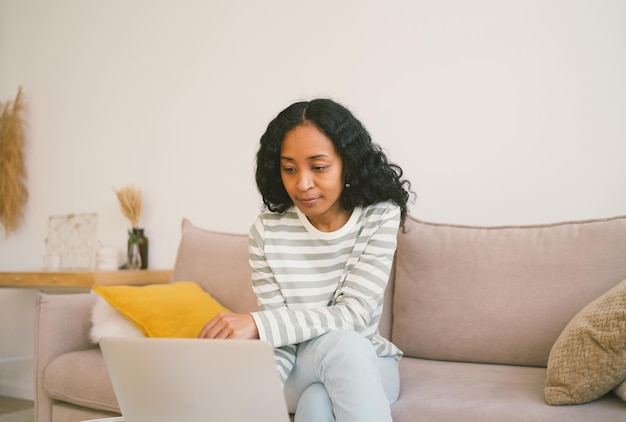 Africanamerican female browsing laptop while sitting on sofa using digital device for work and study