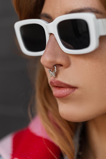 Amazing street closeup portrait of pretty fashion woman with stylish white sunglasses with noise piercing in fashionable urban clothes on a dark background Female hipster face