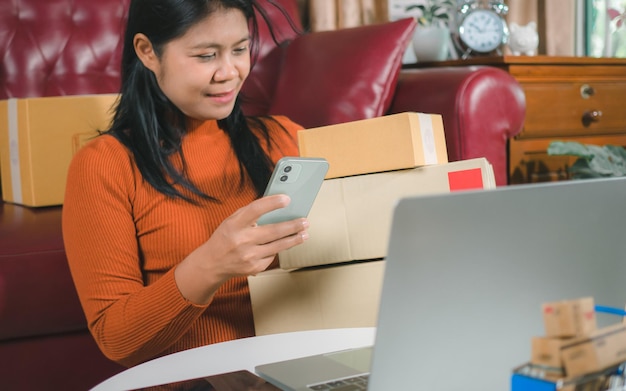 Photo asia women working business sme online at home her checking order from smartphone for customer and online delivery for ready packing checking orders from customers with digital laptop