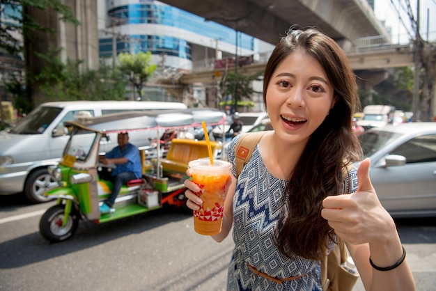 Photo asian backpacker gives a big thumb to the local milk tea holding in her hand. she looks at the camera and praises it.