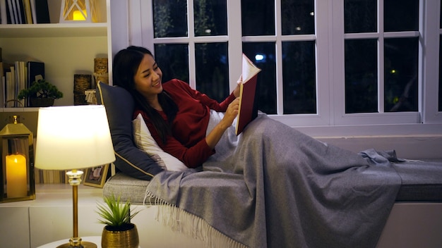 Asian woman reading book in living room at night. Lifestyle and relaxing at home.