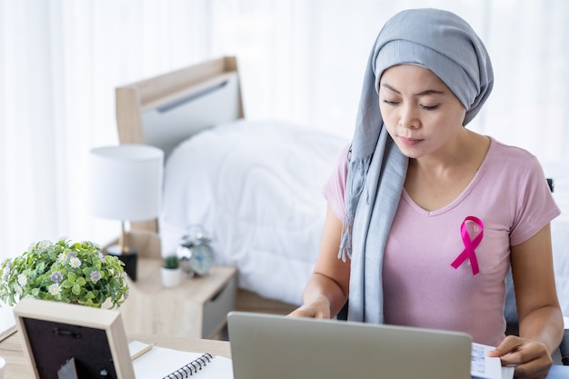 Photo a asian women disease mammary cancer patient with pink ribbon wearing headscarf after treatment to chemotherapy with working business at laptop in office at home,medicine concept