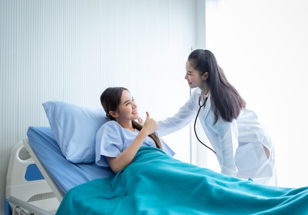 Photo asian young female patient on bed showing thumbs up with smiley face very good symptom to asian young female doctor in hospital background.