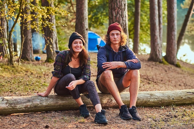 Attractive happy couple is relaxing in the green sunny forest while sitting on the log. There are campsite at background.