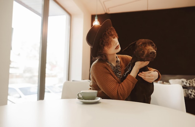 Attractive hipster girl in trendy clothes sits at a table in a coffee shop with a dog in her arms