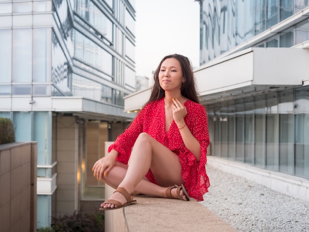 An attractive smiling Asian girl in red sits on a granite dais among glass buildings.