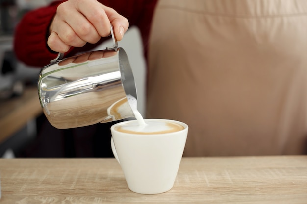 Barista pours milk from metal pot to white glass cup on wooden table.Prepait latte