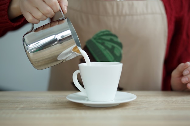 Barista pours milke from steel pot to white cup with coffee