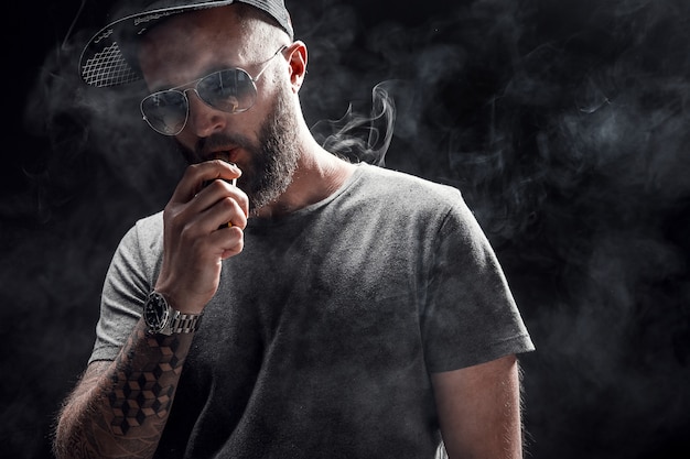 Photo bearded male dressed in a grey shirt, sunglasses and baseball cap vaping