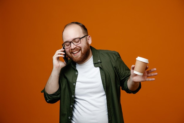 Bearded man in casual clothes wearing glasses talking on mobile phone holding cup of coffee smiling cheerfully standing over orange background