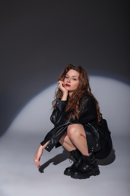 Beautiful fashion stylish woman with a fashionable black leather jacket with a sexy black dress with boots sits on the floor in the studio on a dark background