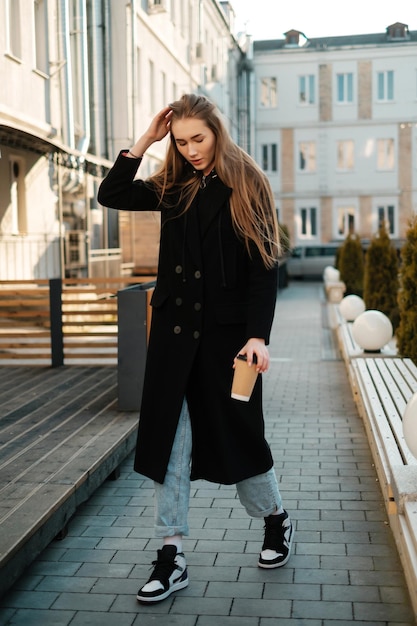 Beautiful girl in a black coat on a walk with coffee to go Casual street female portrait