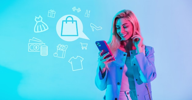 Beautiful happy smiling woman in fashionable clothes holding smartphone and shopping online with shopping icons on blue background. Girl makes purchases on the Internet through the application